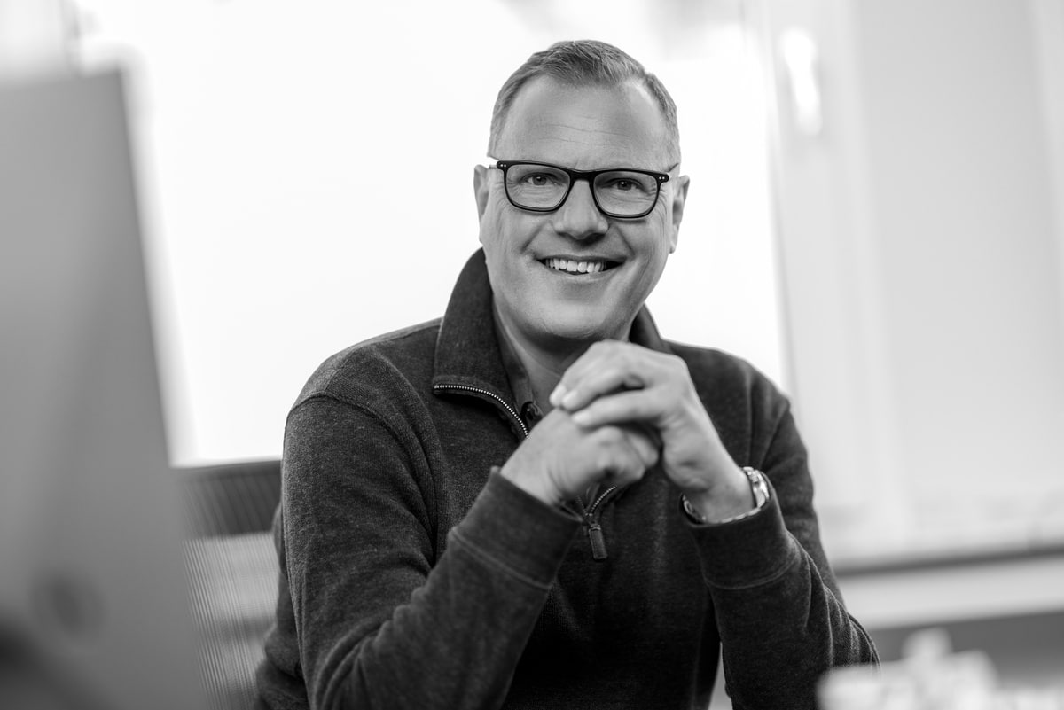 Lächelnder Orthopäde mit Brille im Sprechzimmer, Schwarz-Weiß-Porträt mit gefalteten Händen